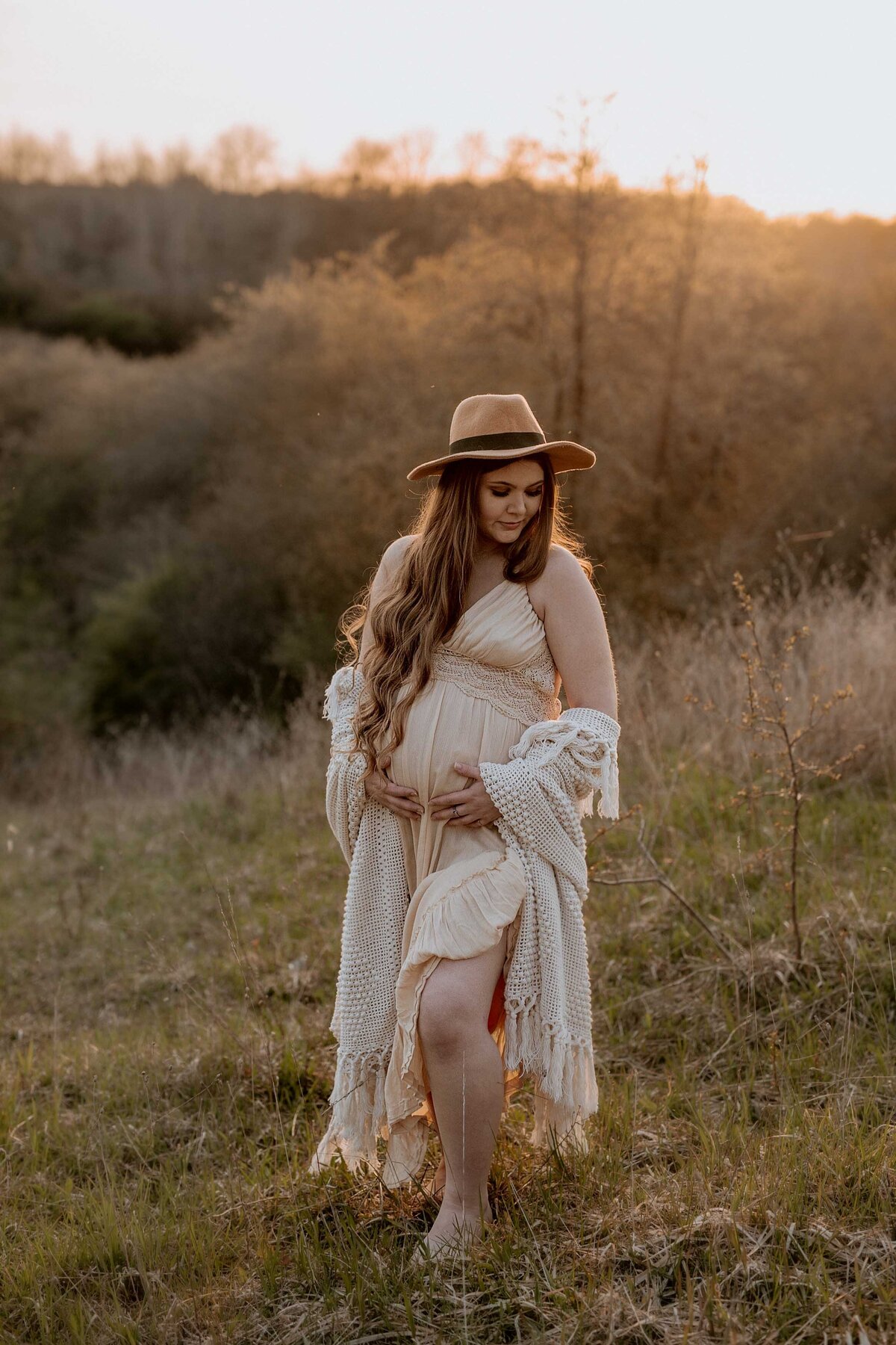 Expectant mom in Exeter, Ontario outdoor maternity photoshoot. Mom is standing barefoot on the grasses in a cream dress and camel felt hat. She is touching her bump and looking down over her shoulder with sun haze in the background.
