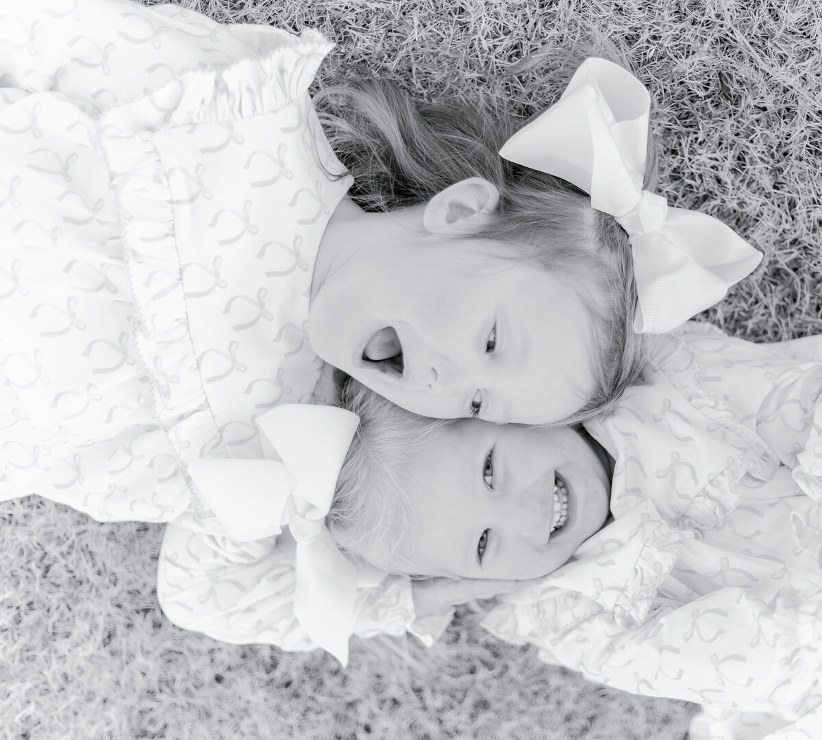 Black and white portrait of two young sisters lying in the grass, dressed in matching ruffled outfits with large white bows. One laughs playfully while the other smiles brightly, capturing the joy and innocence of childhood in a Birmingham family photography session