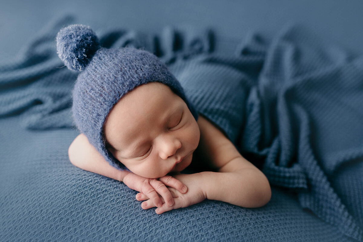 baby boy with bonnet on blue, head on hands pose