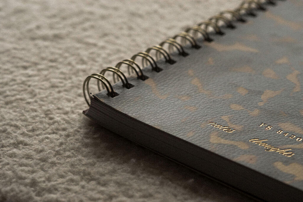 A close-up of a spiral-bound notebook with a patterned cover from Sweet Pea Ink Creative lies on a textured surface, showing the words "notes" and "thoughts" in gold text—perfect inspiration for any brand strategist or web design project.