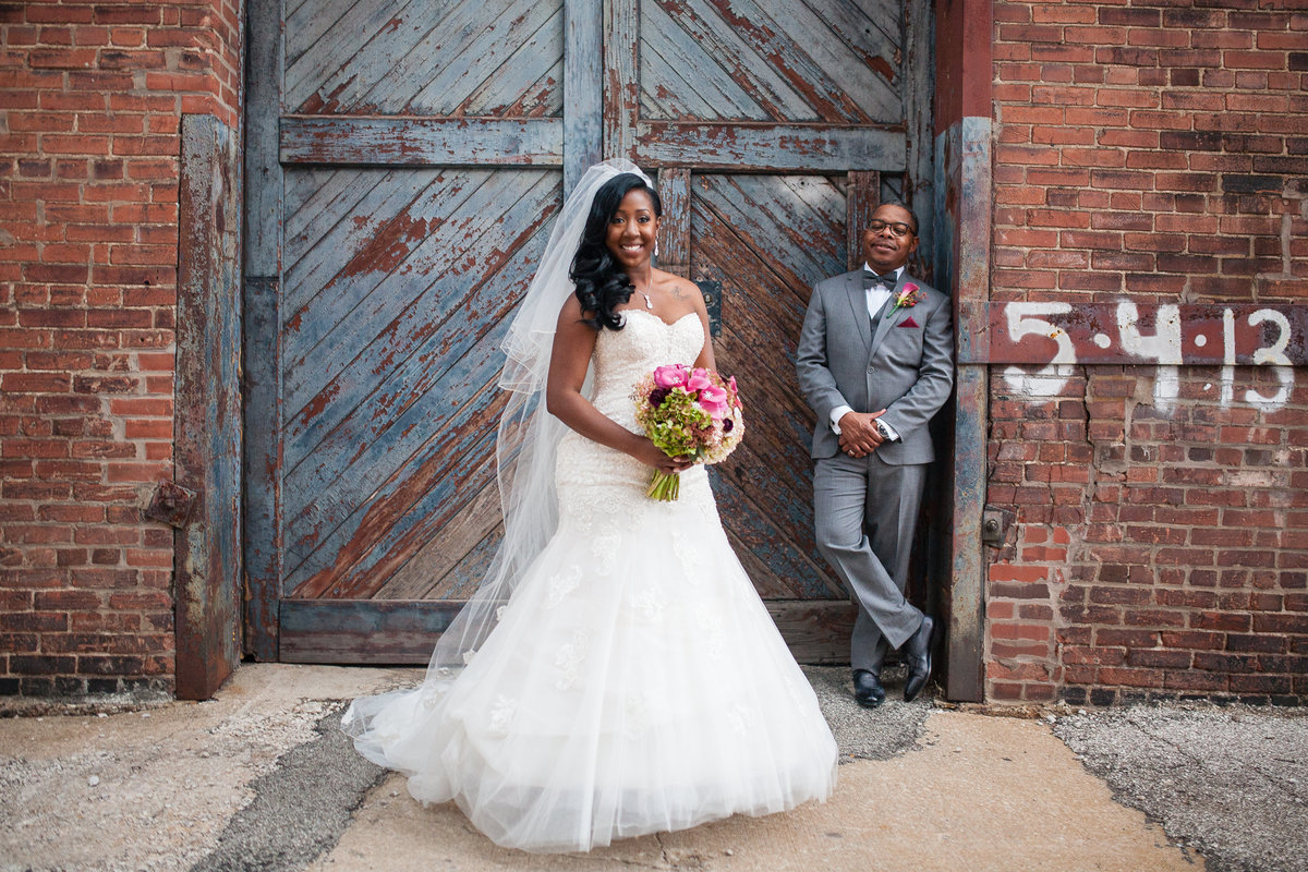 Bride and groom pose in front of wooden doors in St. Charles, the bride in the foreground, as the groom, leaning against a brick wall, admires his new wife.