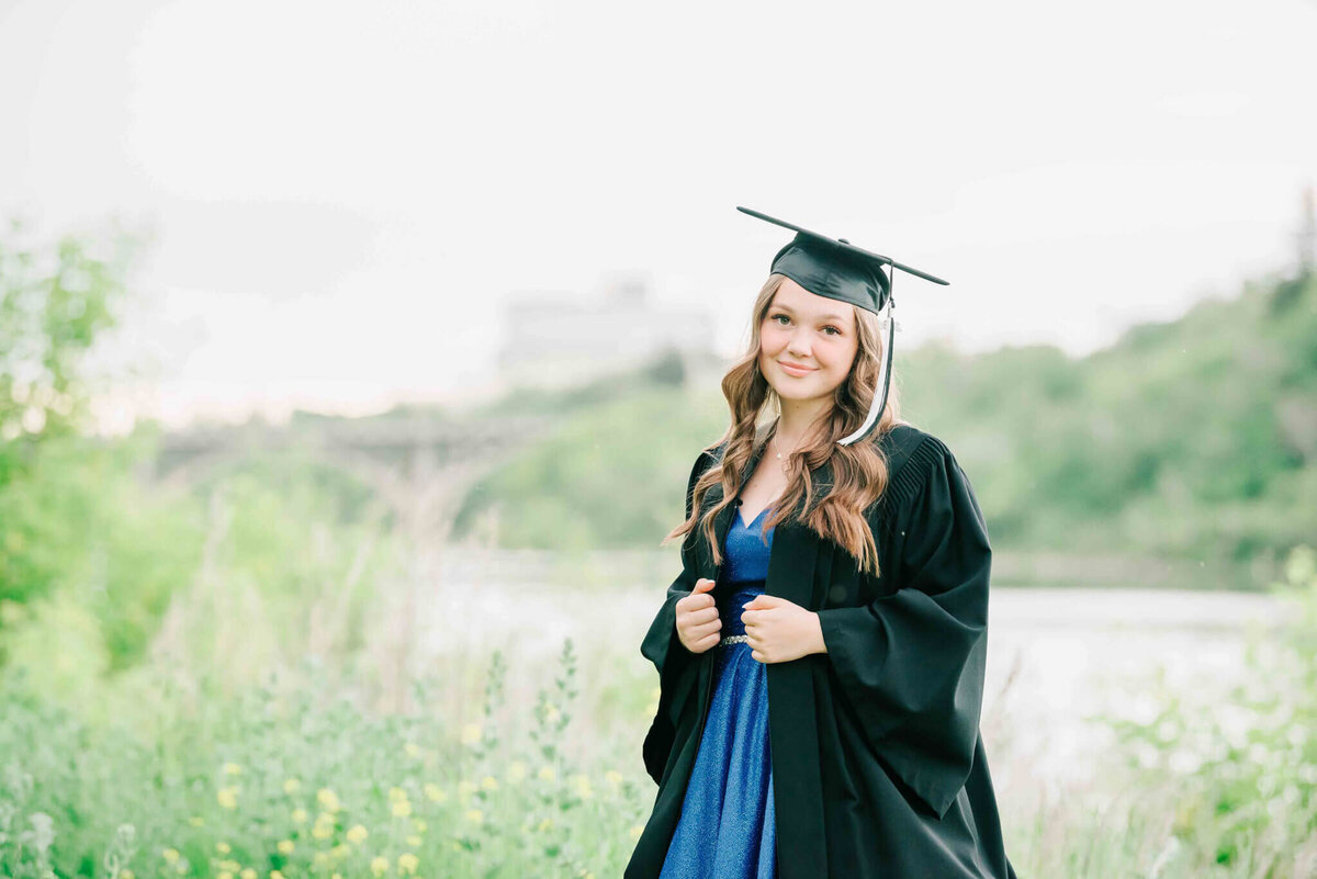 Standing by the river wearing grad cap and gown. Holding onto the front of the grad gown and keeping it open. 