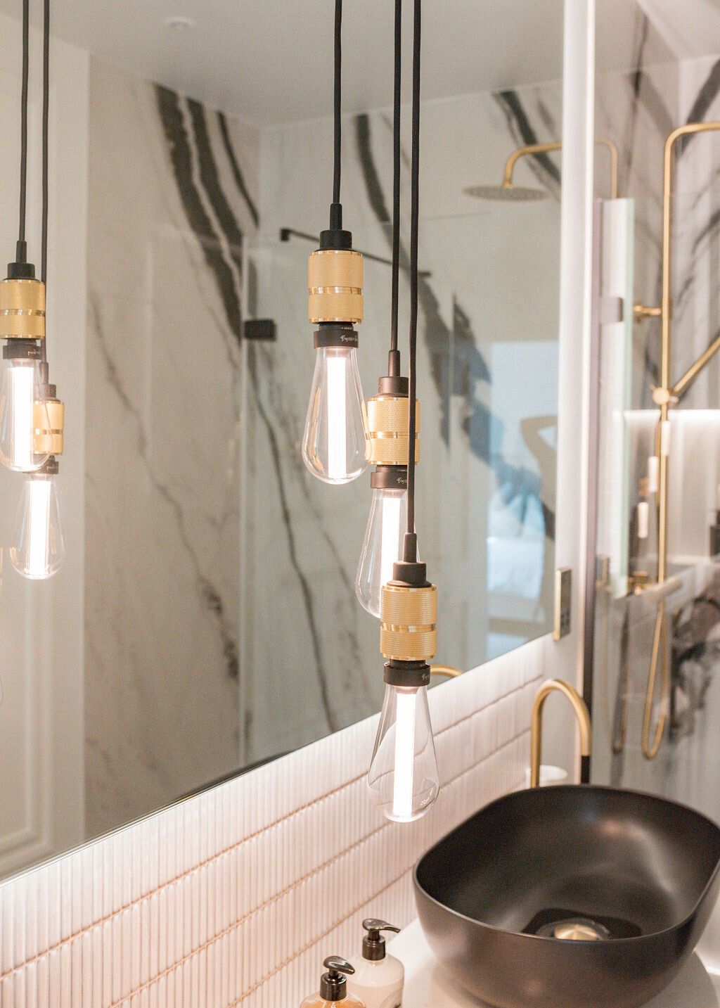 Modern bathroom with marble walls and countertops featuring black veining. A black vessel sink sits on a floating countertop with wooden storage below. Pendant lights hang above the sink, and a glass-enclosed shower with gold fixtures is visible in the background.