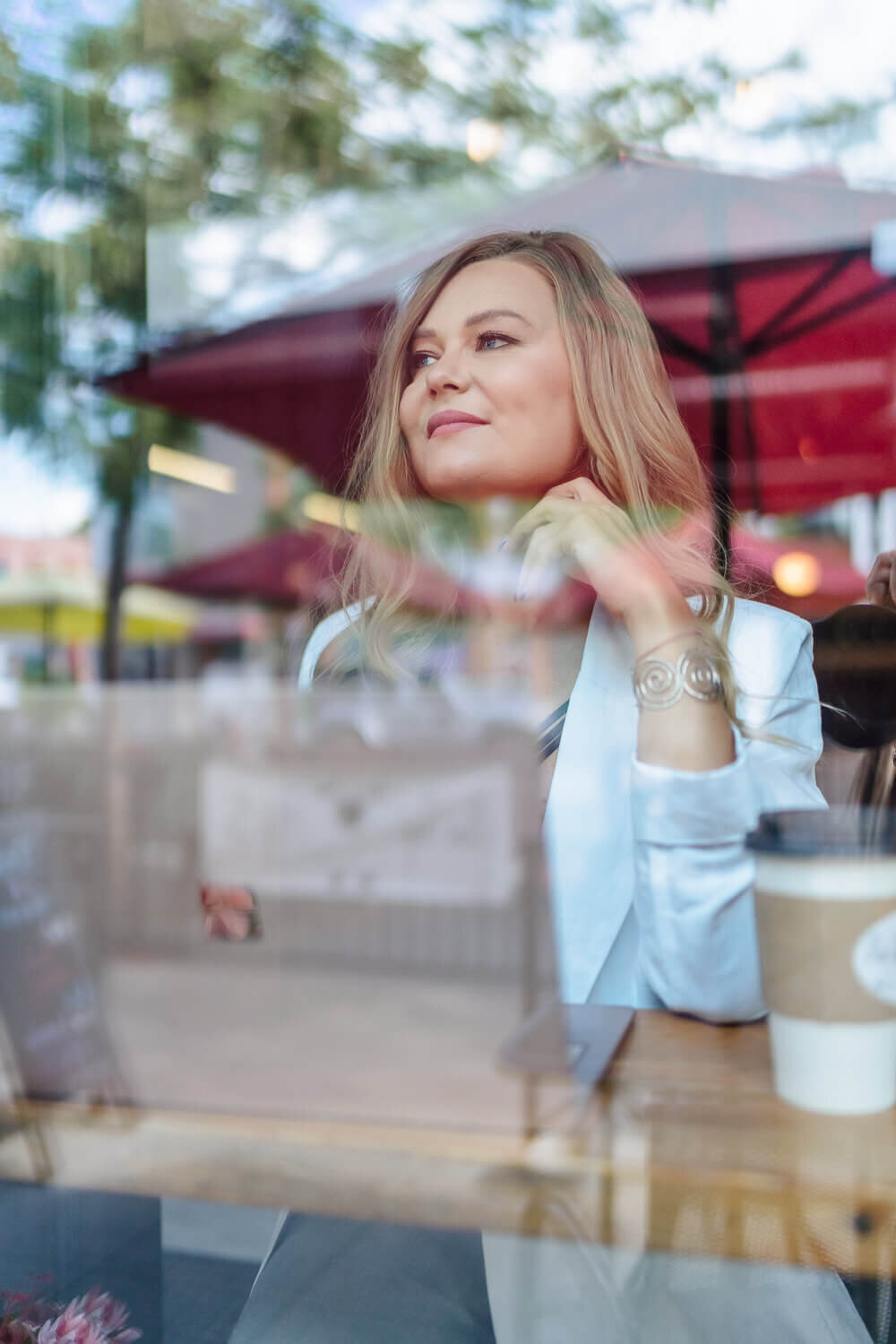 Female entrepreneur seen through café window holding coffee cup, smiling while looking outside.