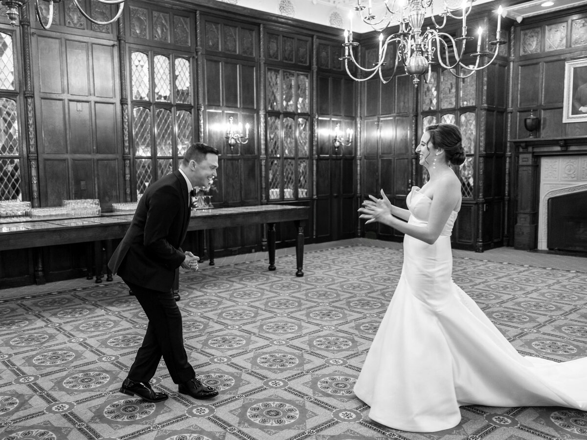 A bride in a strapless gown and a groom in a suit share a joyful moment in an elegant room with wood-paneled walls and chandeliers. The groom is leaning forward, smiling, while the bride gestures excitedly.