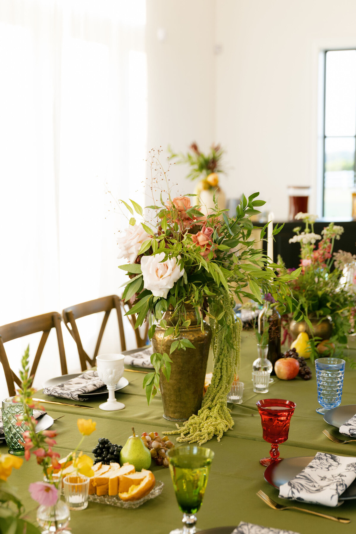 A close-up view of a long wedding dining table decorated with lush garden-inspired centerpieces featuring roses, greenery, pops of color, fruit details, taper candles, and vintage glassware, styled by a wedding florist for a colorful reception design.