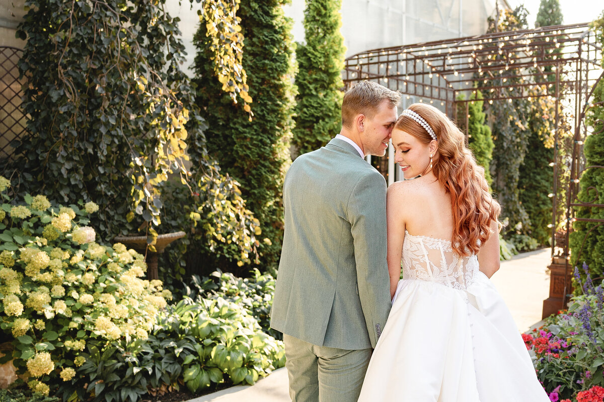 A newlywed couple stands in a beautifully landscaped garden, with the groom in a sage green suit and the bride in a white wedding gown with lace details. They are smiling and gazing at each other lovingly, surrounded by greenery and flowers.