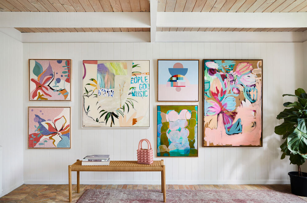 A gallery wall in a living room with several abstract paintings in shades of pink, blue, green, and orange. The room has white paneled walls and a light wood ceiling.