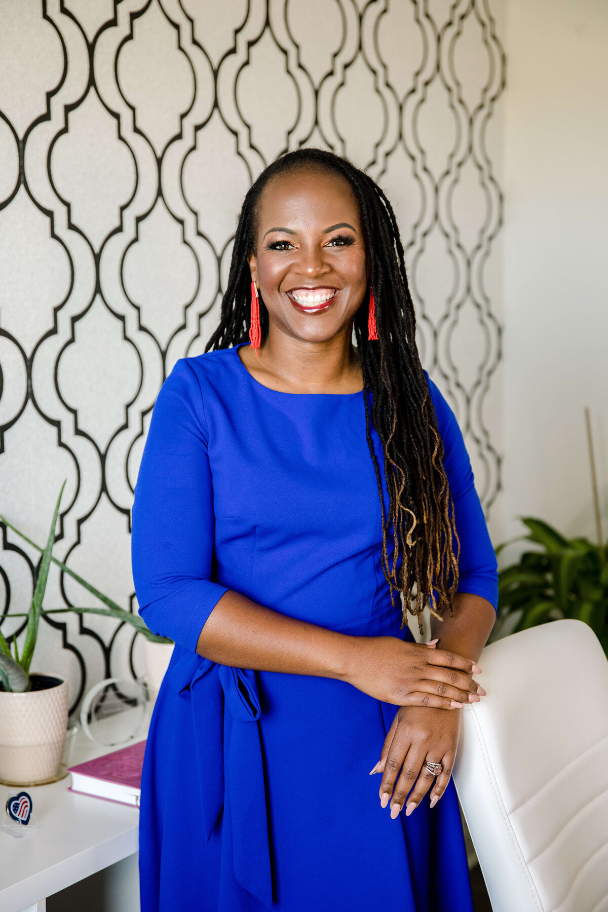 headshot of black business owner in a dark blue dress posing in her home office and smiling as she holds her hands together captured by commercial photographers