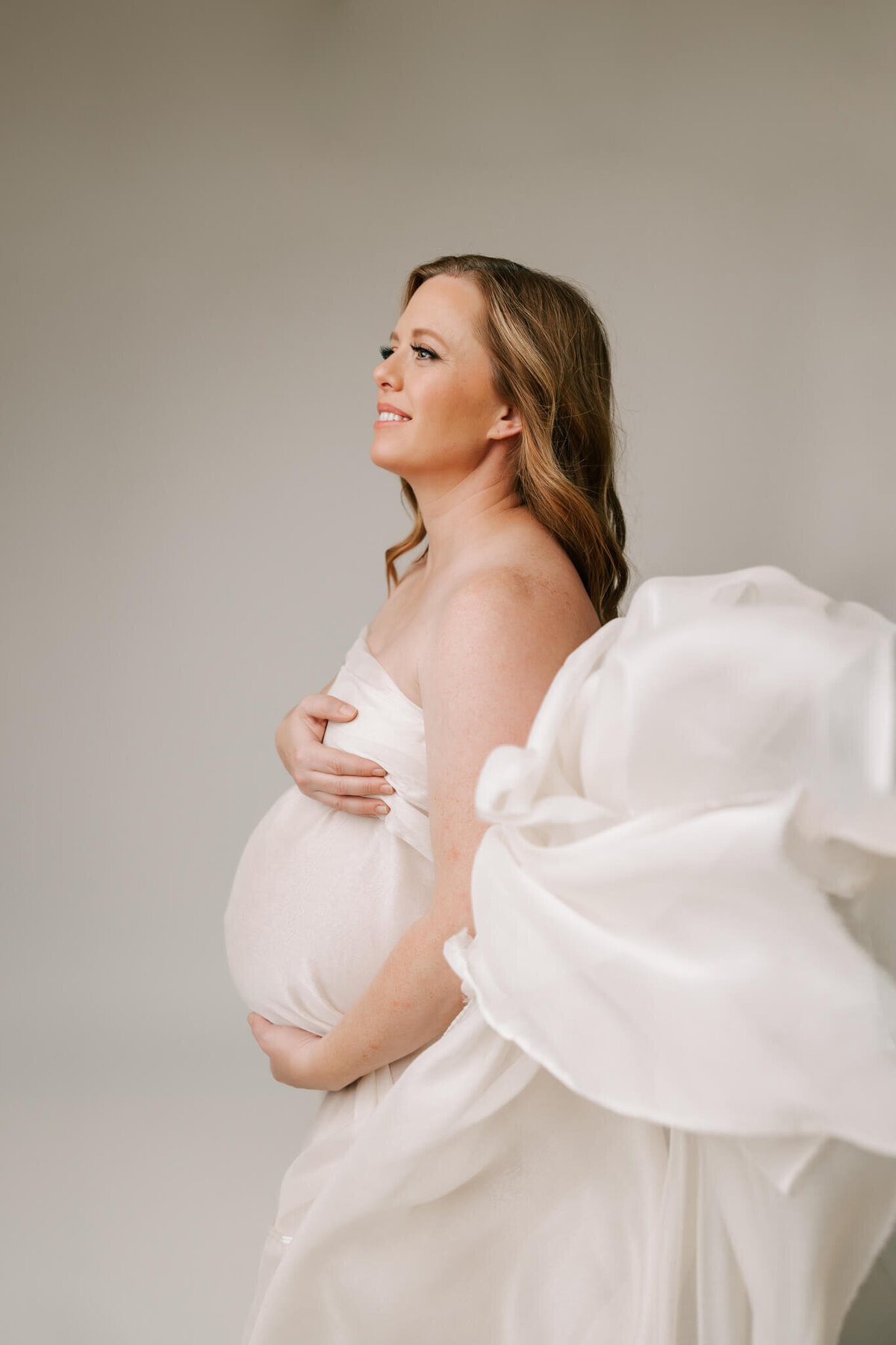 red-headed woman draped in cream silk smiling off to the side while fabric flows around her in studio photography session.