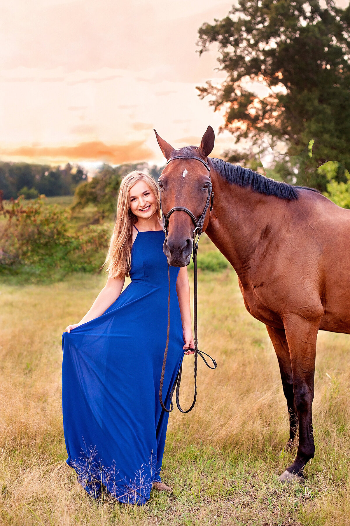 Clover Hill high school senior girl wearing a long blue dress smiles with her horse in a field at sunset for her Richmond, VA senior portraits.
