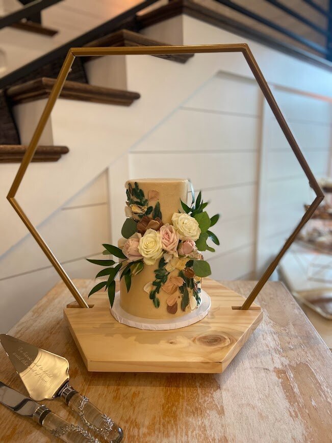 A two tier cutting cake sits on a wooden cake stand. Across the front are painted buttercream flowers and leaves going in one direction, and in the other direction across the front are fresh roses and greenery.
