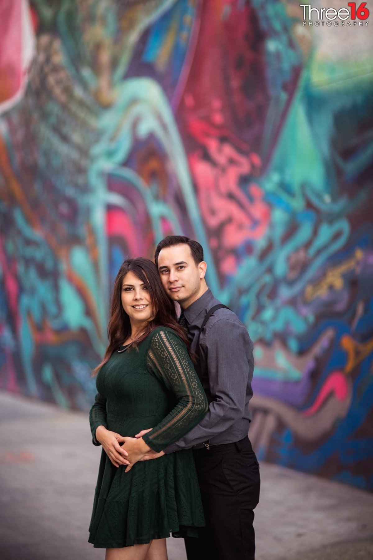 Groom to be embraces his fiance from behind in front of a colorful mural on a wall