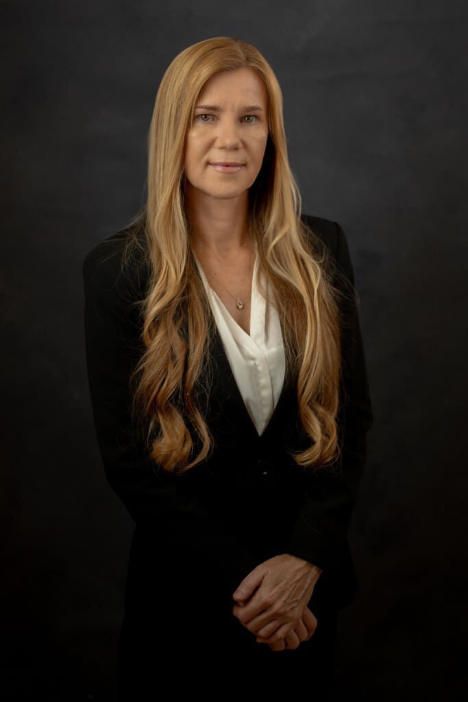 Professional-woman-with-long-blonde-hair-in-a-black-suit-jacket-and-white-blouse-posing-against-a-dark-background