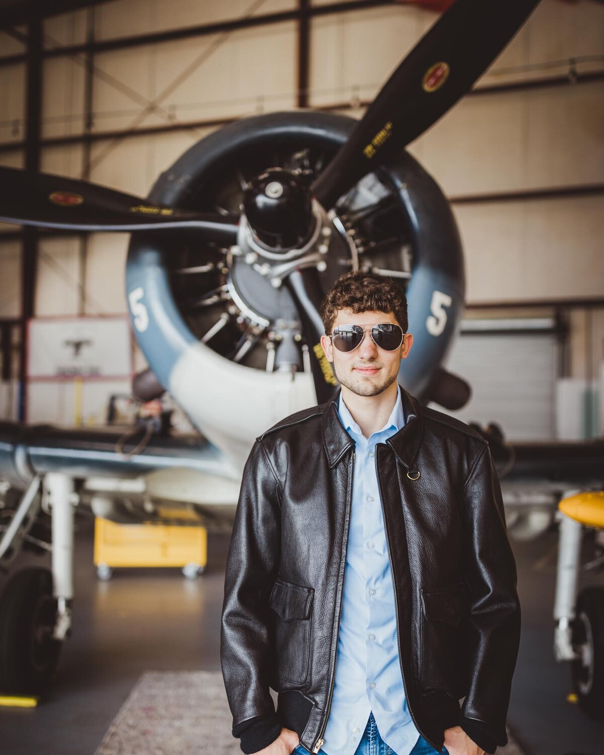Future pilot in front of an airplane in a hanger with his leather jacket and aviator glasses on.