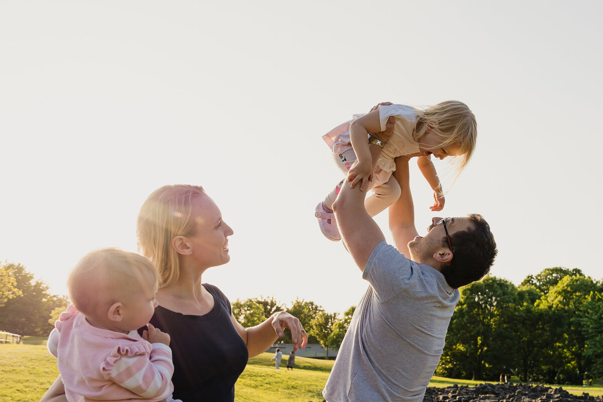 A joyful family outdoors at sunset, with a father lifting a young girl high in the air and a mother holding a baby.