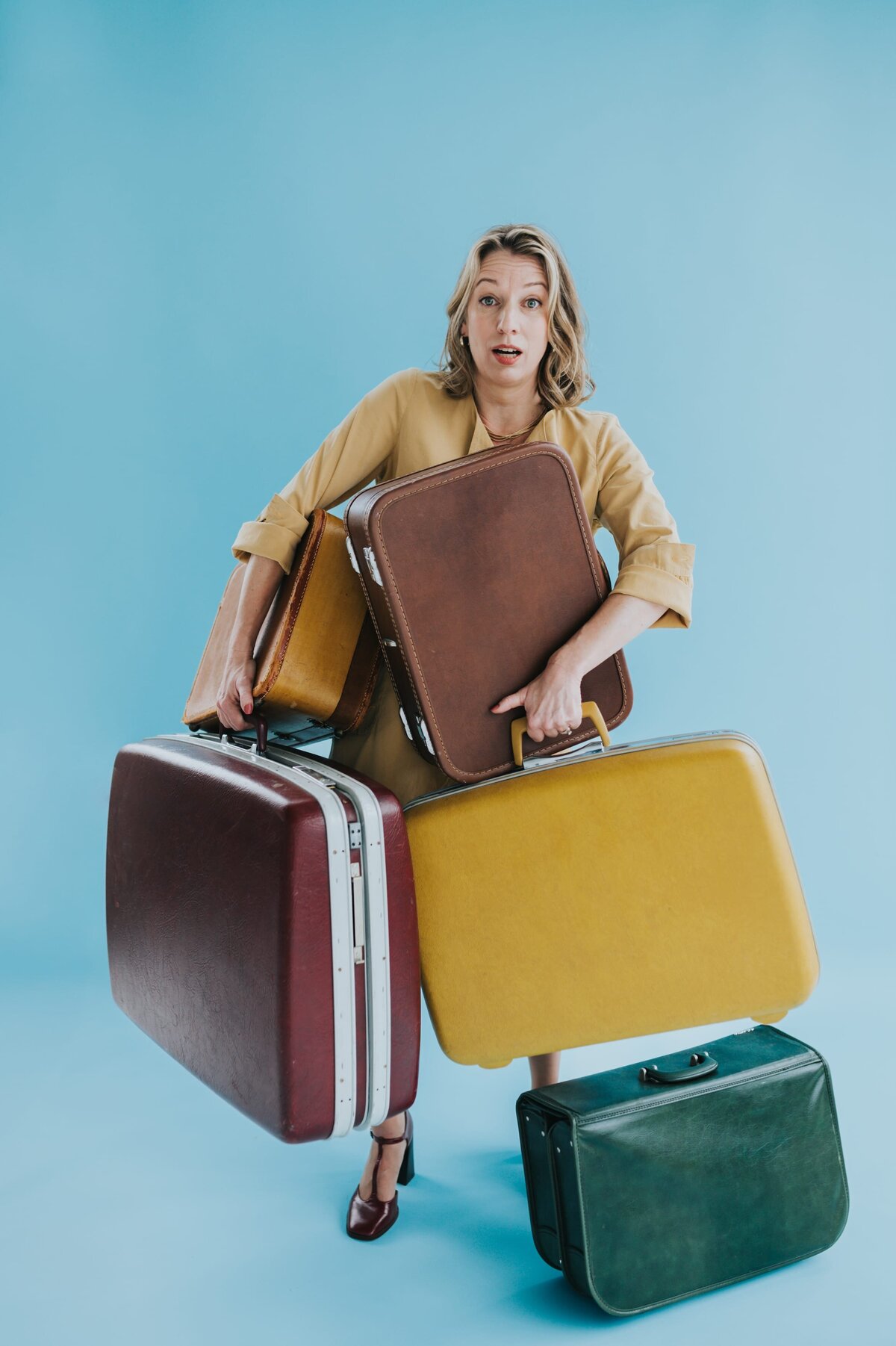 Woman with shoulder-length blonde hair holds several vintage suitcases at once with a shocked expression on her face in front of an aqua backdrop.
