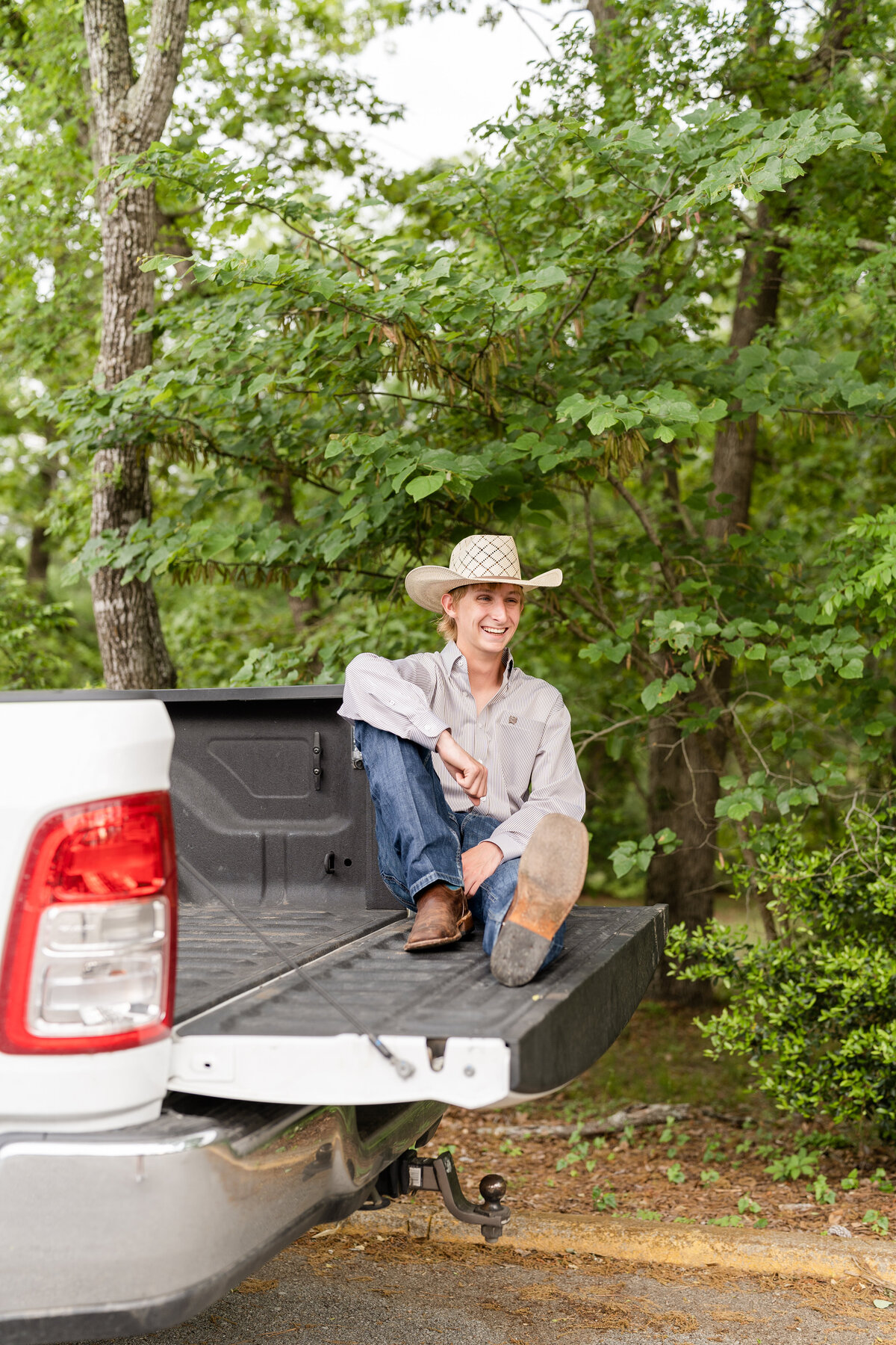 High School senior guy sitting on truck tailgate laughing away with arm on knee  while wearing button down, cowboy hat, cowboy boots and jeans