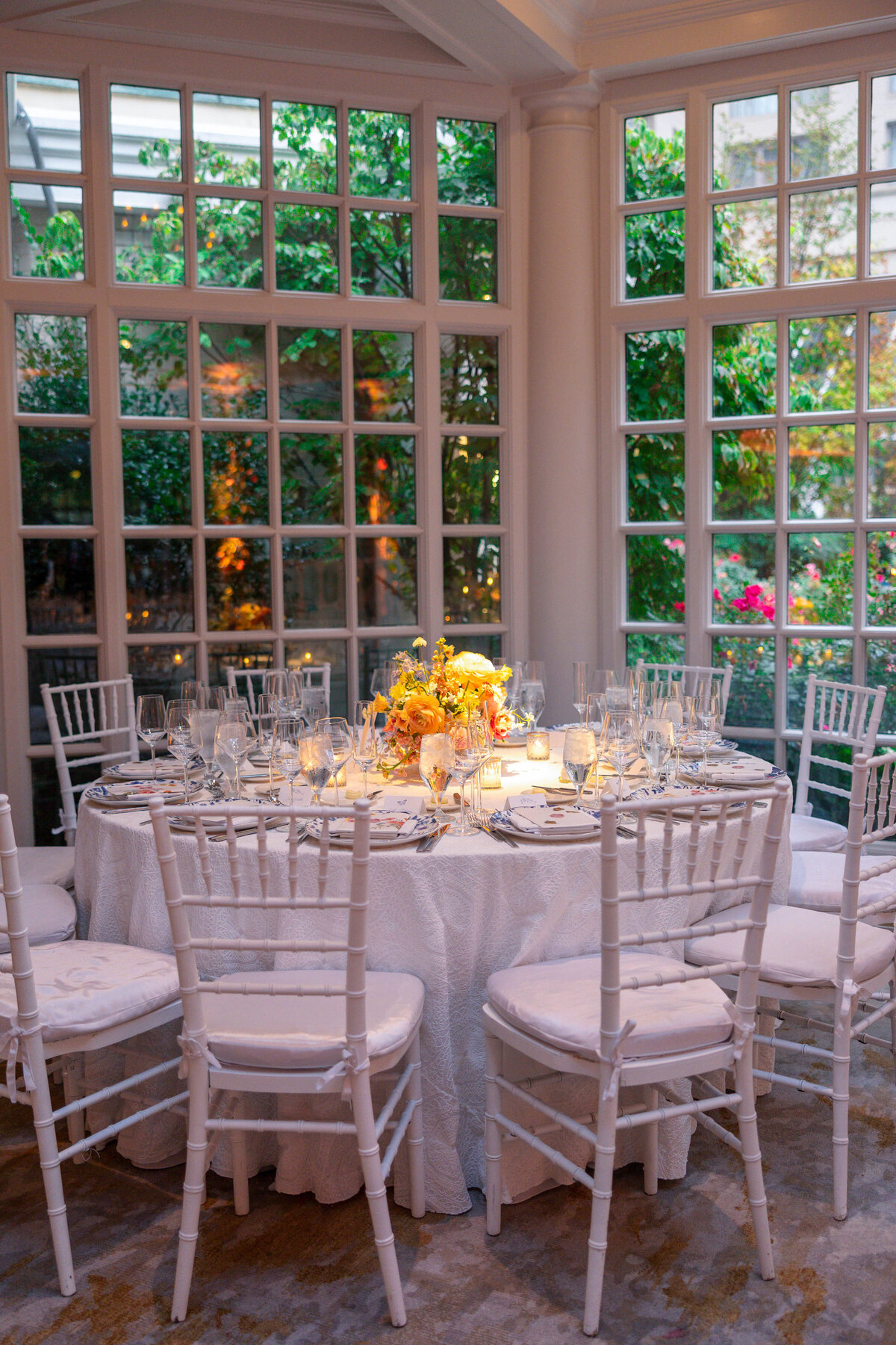 Elegant table setting with white chairs around a round table, adorned with a floral centerpiece and glassware, in a bright room with large windows overlooking a garden.