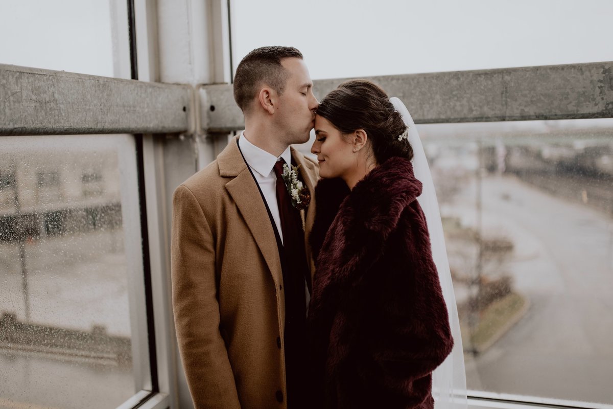 Groom kissing bride's forehead, wearing coats and keeping each other warm inside, next to glass walls.