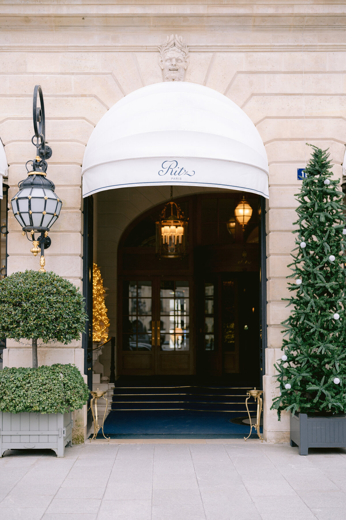 An elegant storefront with a white, rounded awning that reads Ritz above the entrance. Flanked by manicured topiary plants in square planters on the left and a decorated Christmas tree on the right. Ornate lamps hang beside the doorway, and a chandelier is visible inside.