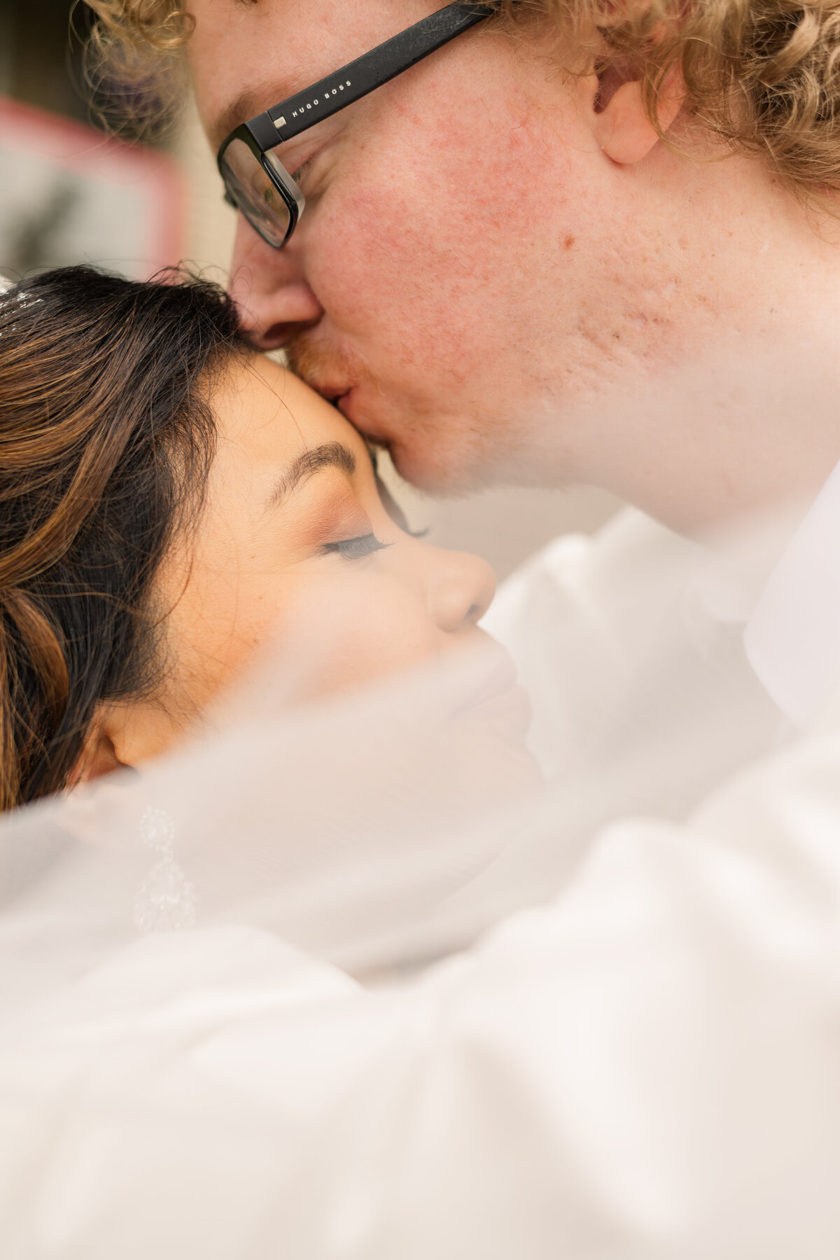 A groom gently kisses the forehead of the bride with the veil softly swished in front of them on their micro wedding day