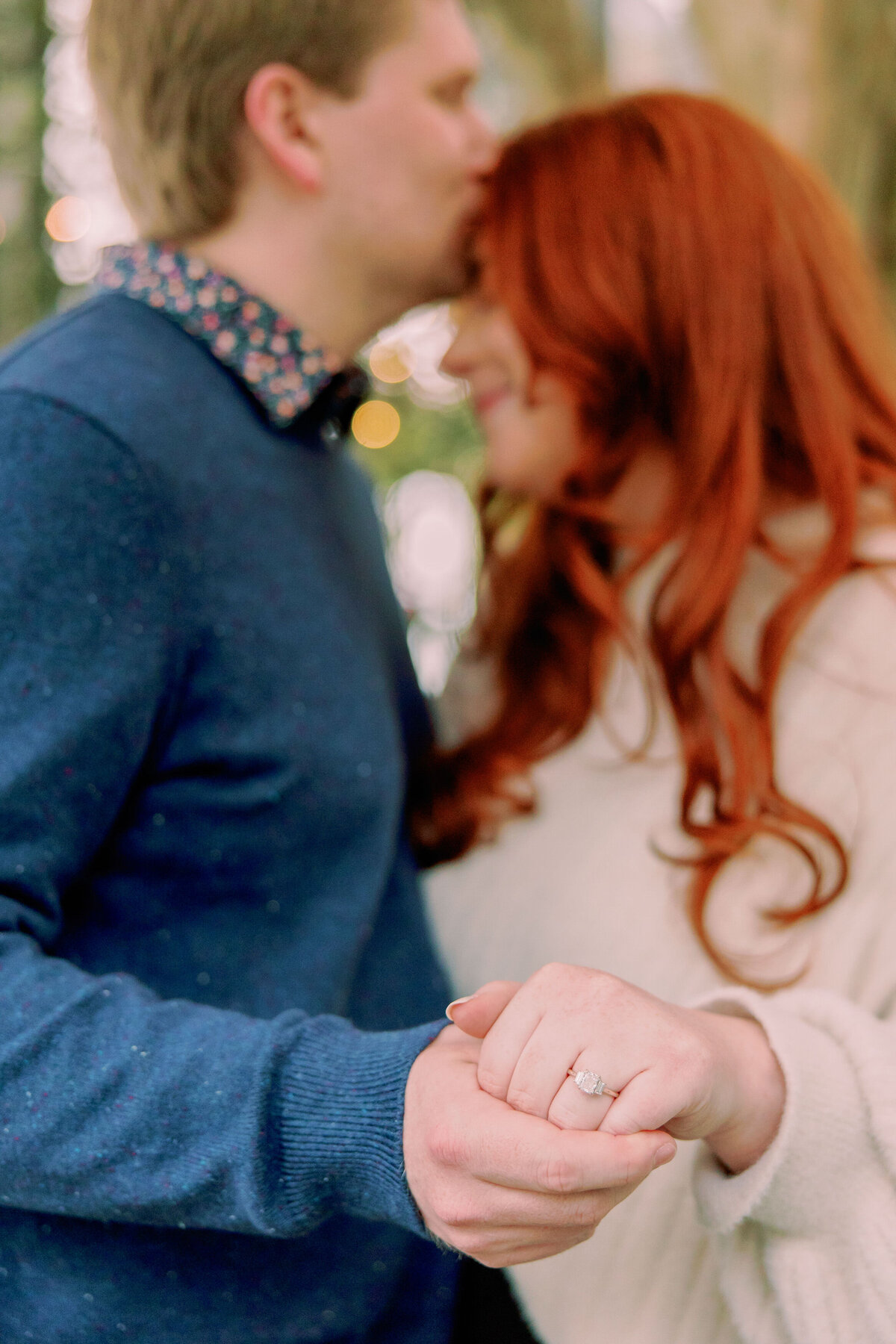 A person kissing their partner's head and holding out their hand to show off an engagement ring 