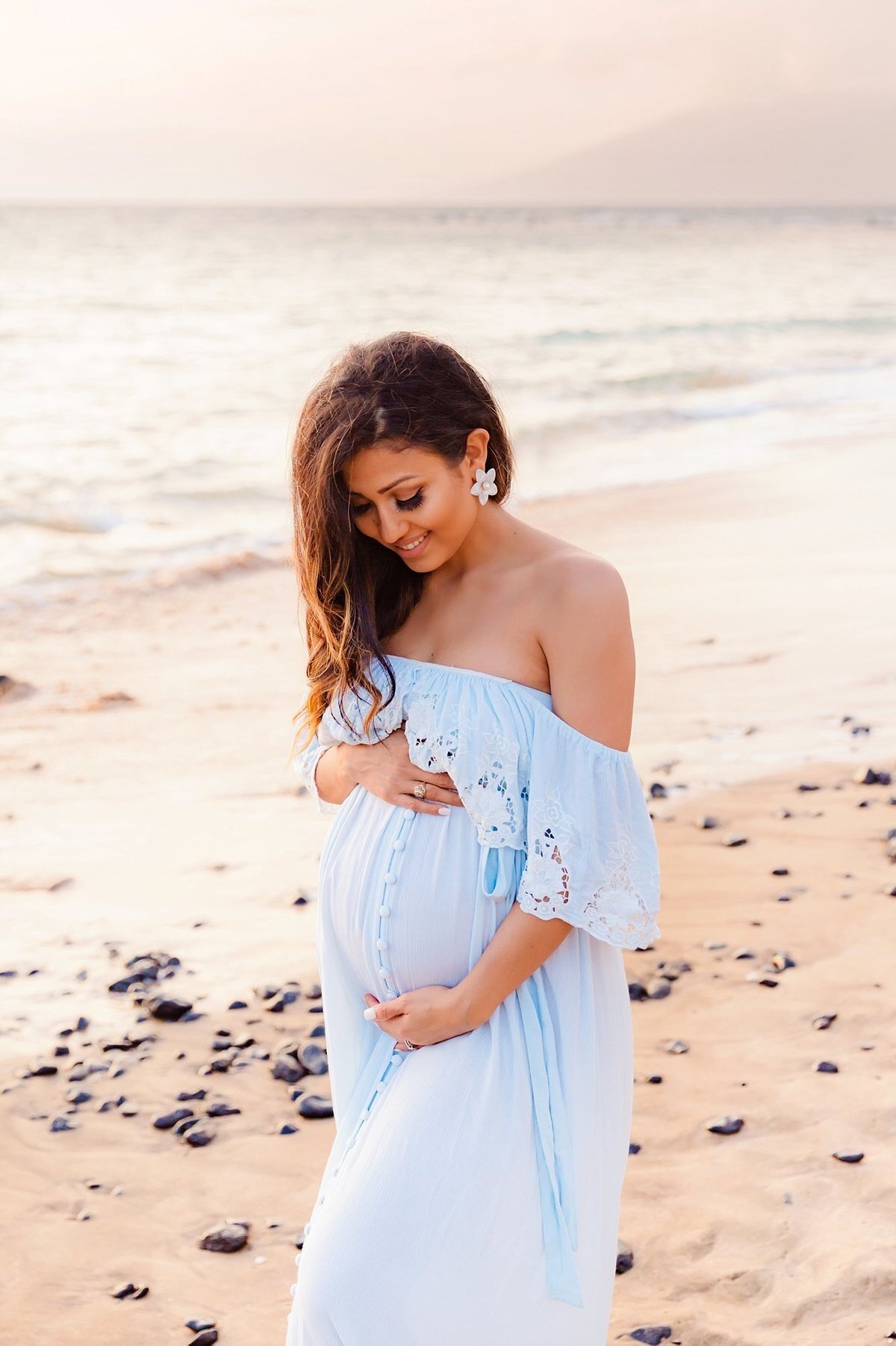 Woman in blue maternity gown with frilly neckline smiles at her pregnant belly during her maternity portraits on Maui