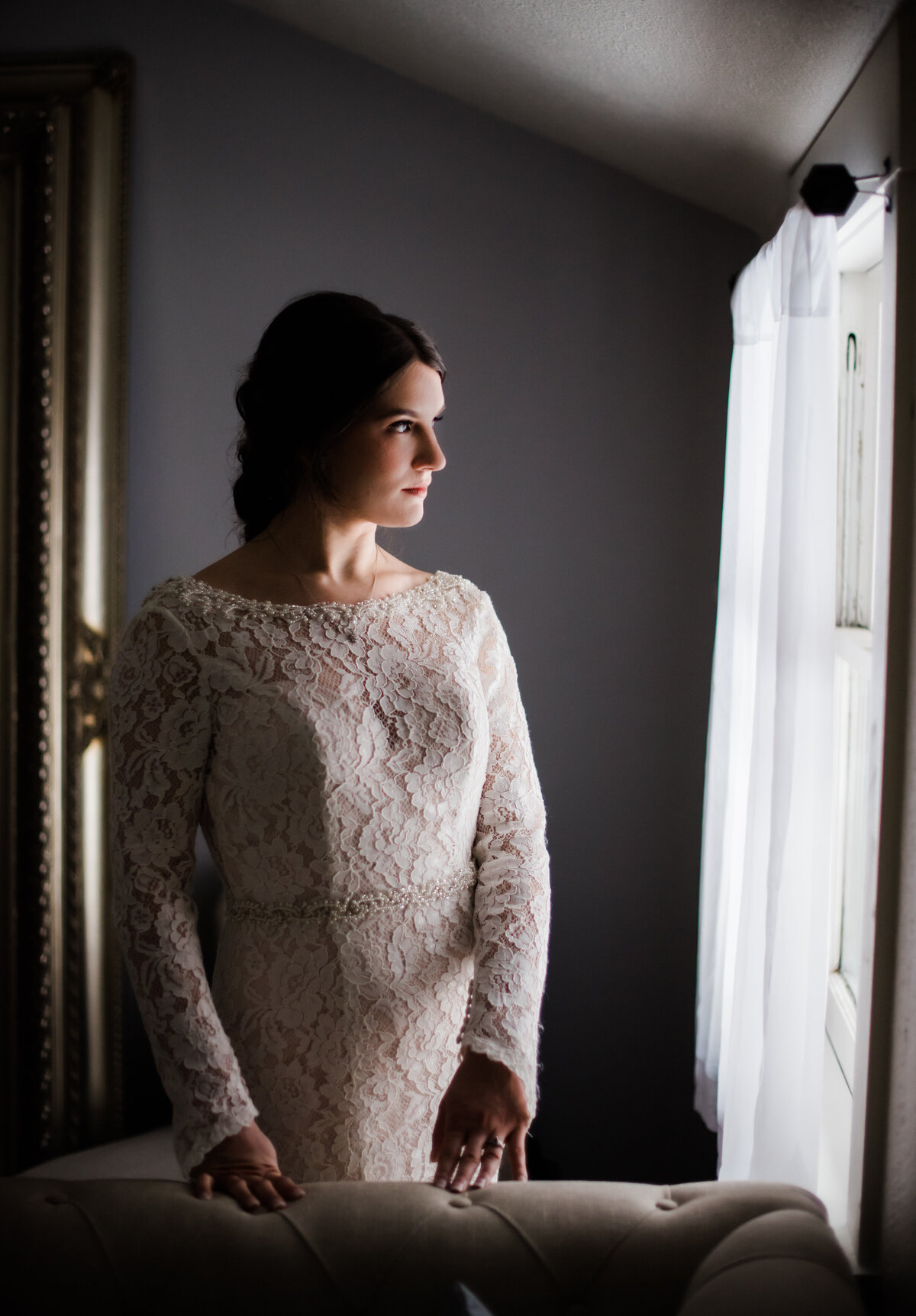 Denver wedding photographer captures bride with her hands resting on a couch in the bridal suite in her wedding dress while looking out the window