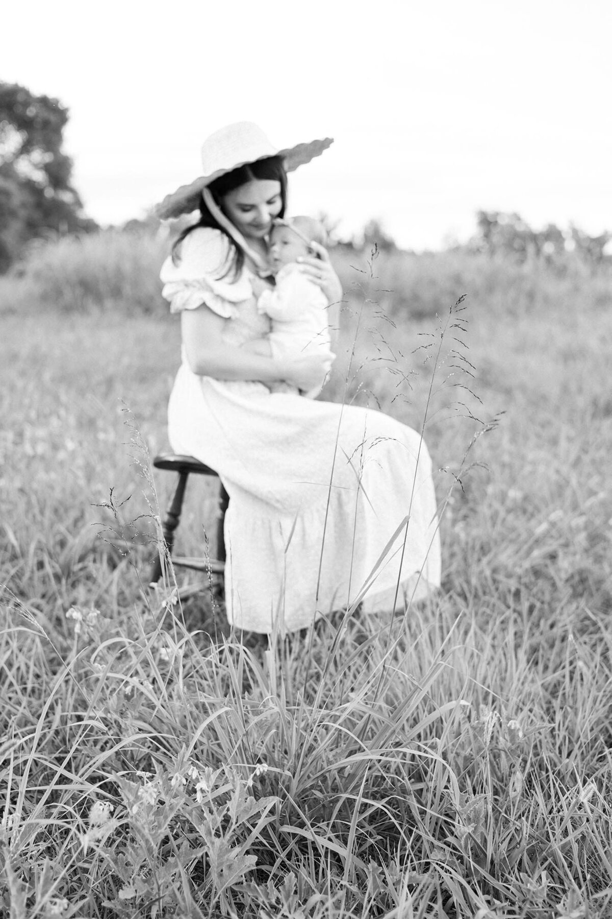 Black and White Virginia BEach Newborn Photography Portrait of a Mother snuggling her newborn close sitting on a stool in a grassy field  by Mary Eleanor