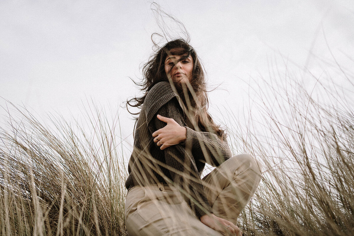vrouw zittend in de duinen met harde wind, donkere kleding, strand portret