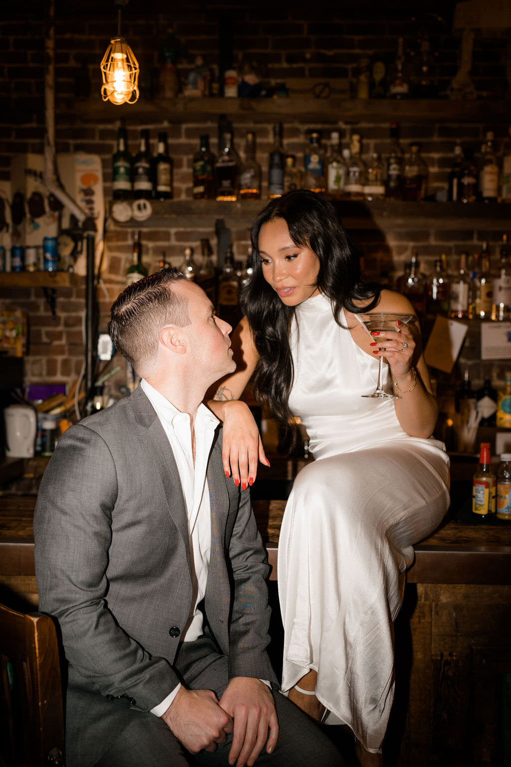 groom sitting in a chair at a bar while his bride sits on the bar and rests her arm on his shoulder