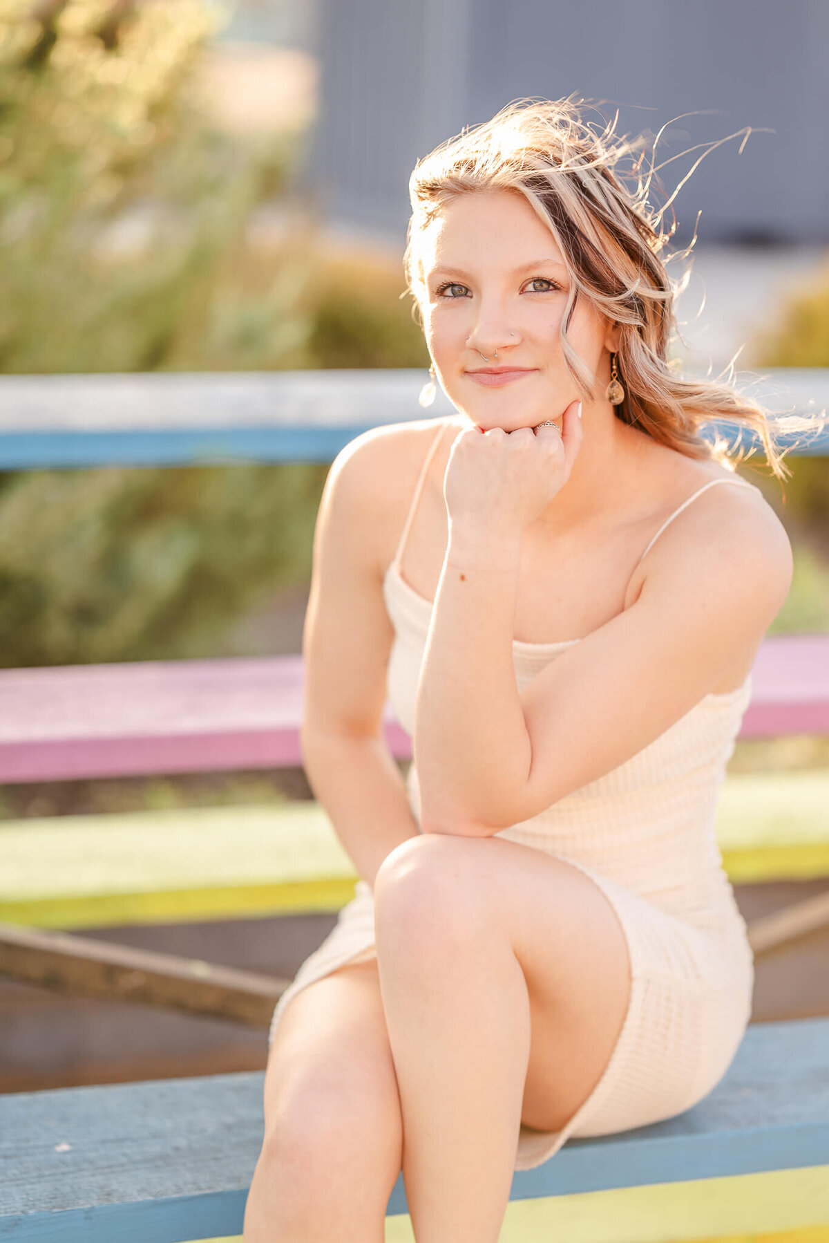 A high school senior in an off-white bodycon dress, sits on some rainbow painted risers. She has her knee propped up and is resting her chin on her fist.