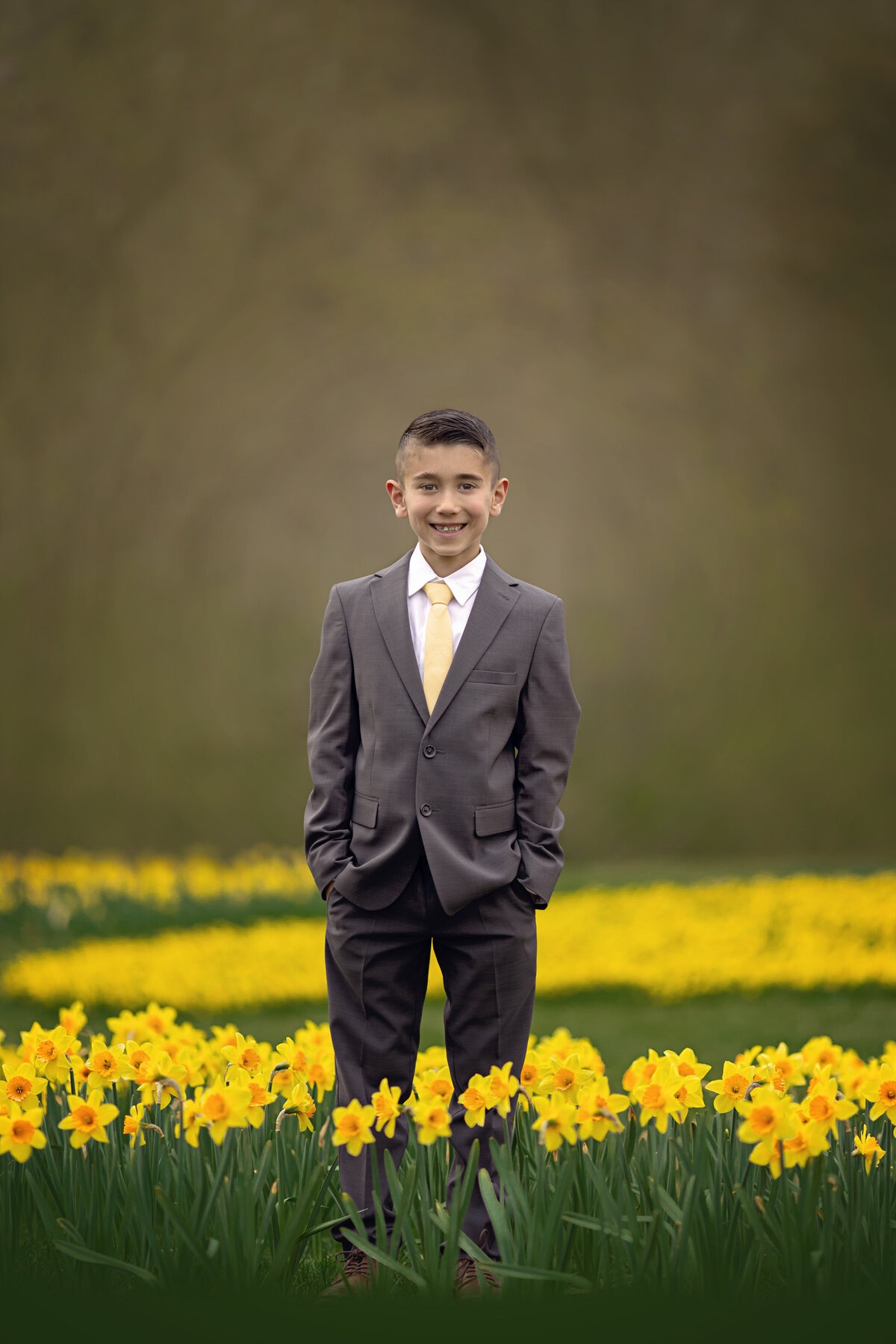 A young boy in a grey suit stands among some yellow daffodils in a grey suit and yellow tie with hands in his pockets