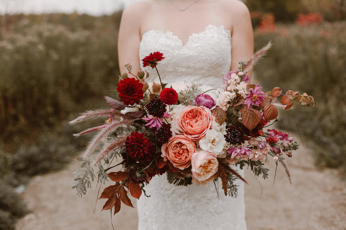 A bride standing in an autumn field holds a bouquet of roses, dahlias and organic greenery. 