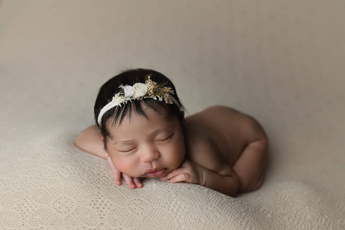 A newborn baby sleeps in froggy pose with a white flower headband