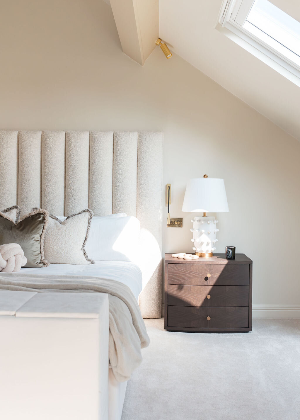 A well-lit bedroom featuring a large plush bed with a tall, tufted headboard. The bed is flanked by a dark wooden nightstand, with a white lamp. The room has a neutral color palette with white and beige bedding and some accent pillows.