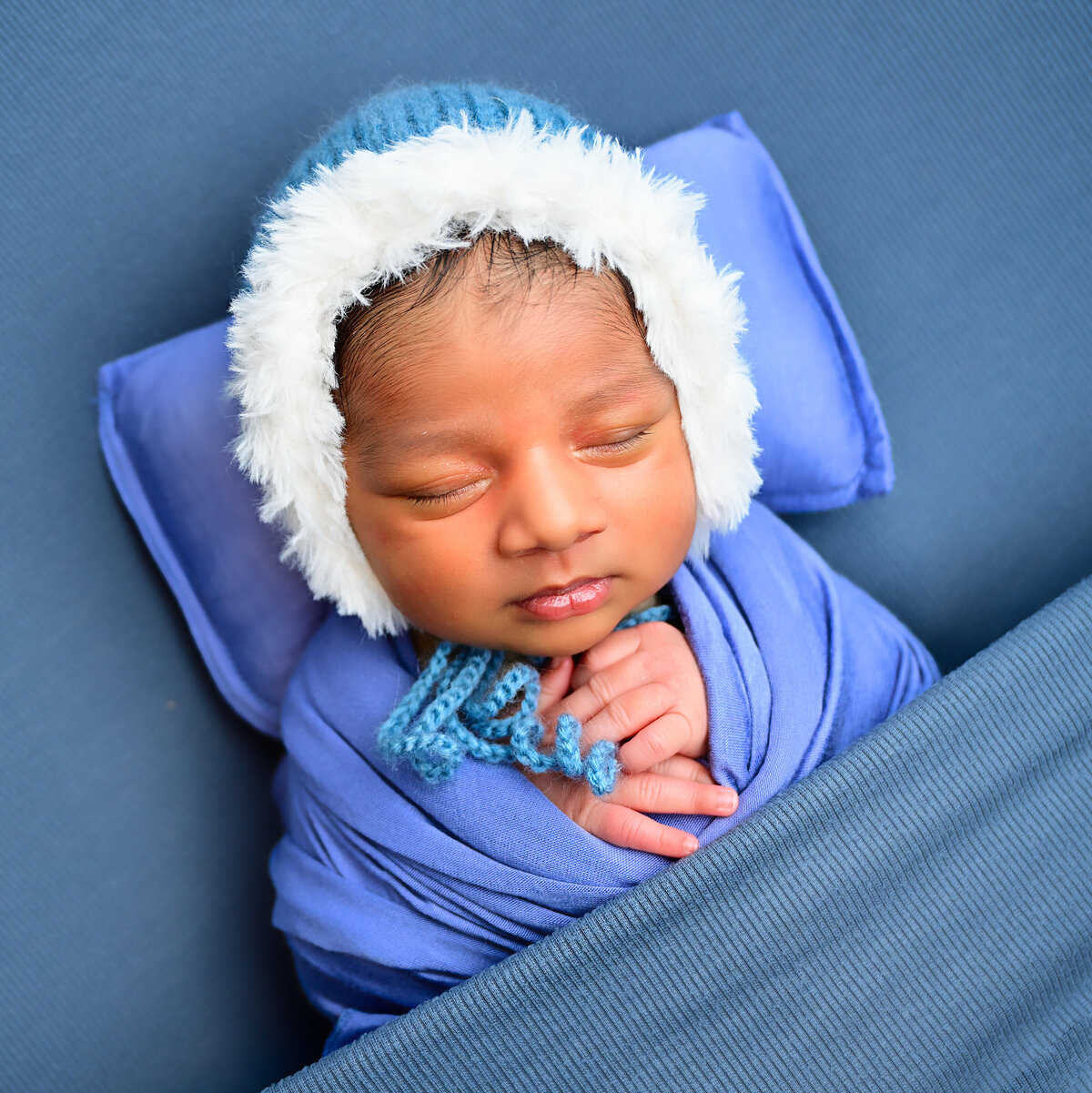Beth Benecki captures this newborn during his newborn photo session. Baby is wrapped in blue and tucked in a blue blanket. Baby is wearing a blue bonnet that has white fur trim.