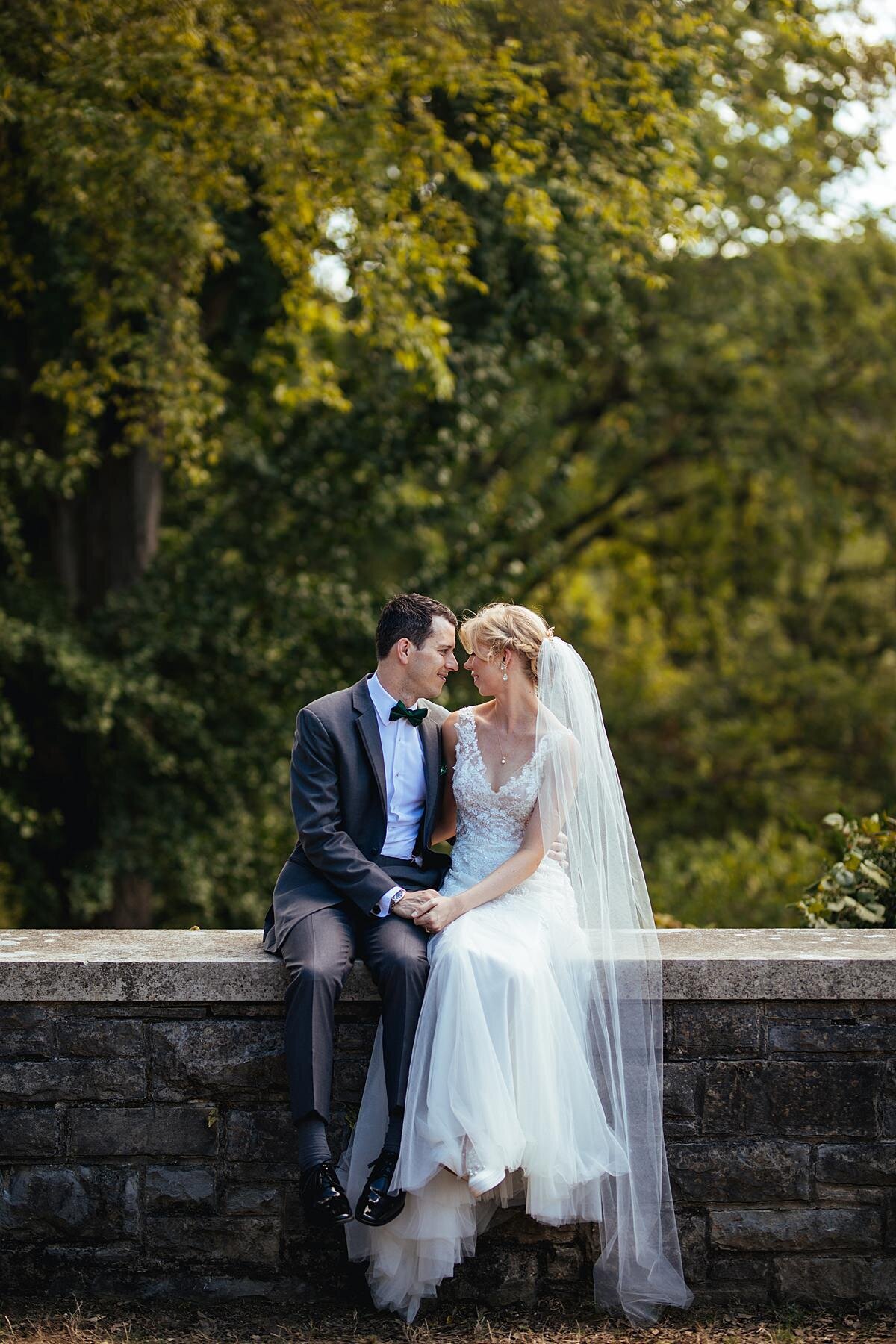 The bride, dressed in a sleeveless lace wedding dress and long veil sits beside the groom in a charcoal gray suit on a low stone wall at Cheekwood holding hands and touching foreheads