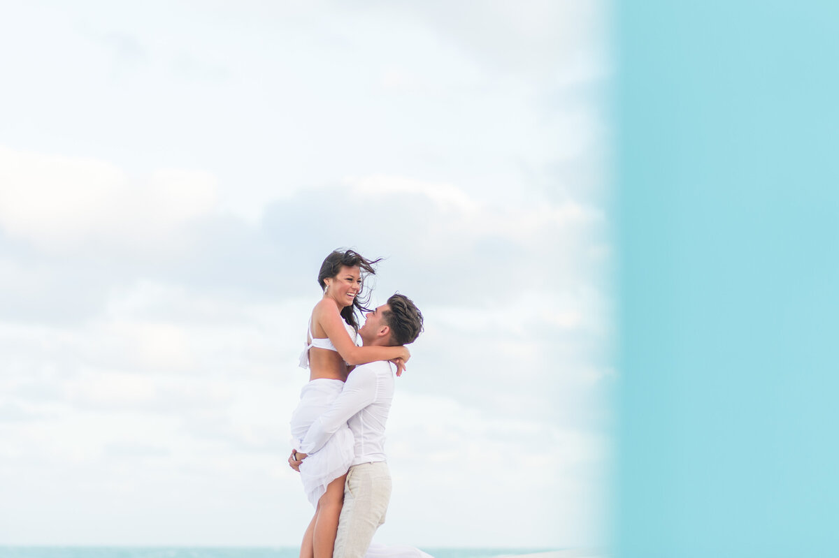 Engaged couple on Miami beach with the man lifting the woman as they share a joyful moment against a cloudy sky, partially framed by a blue wall.