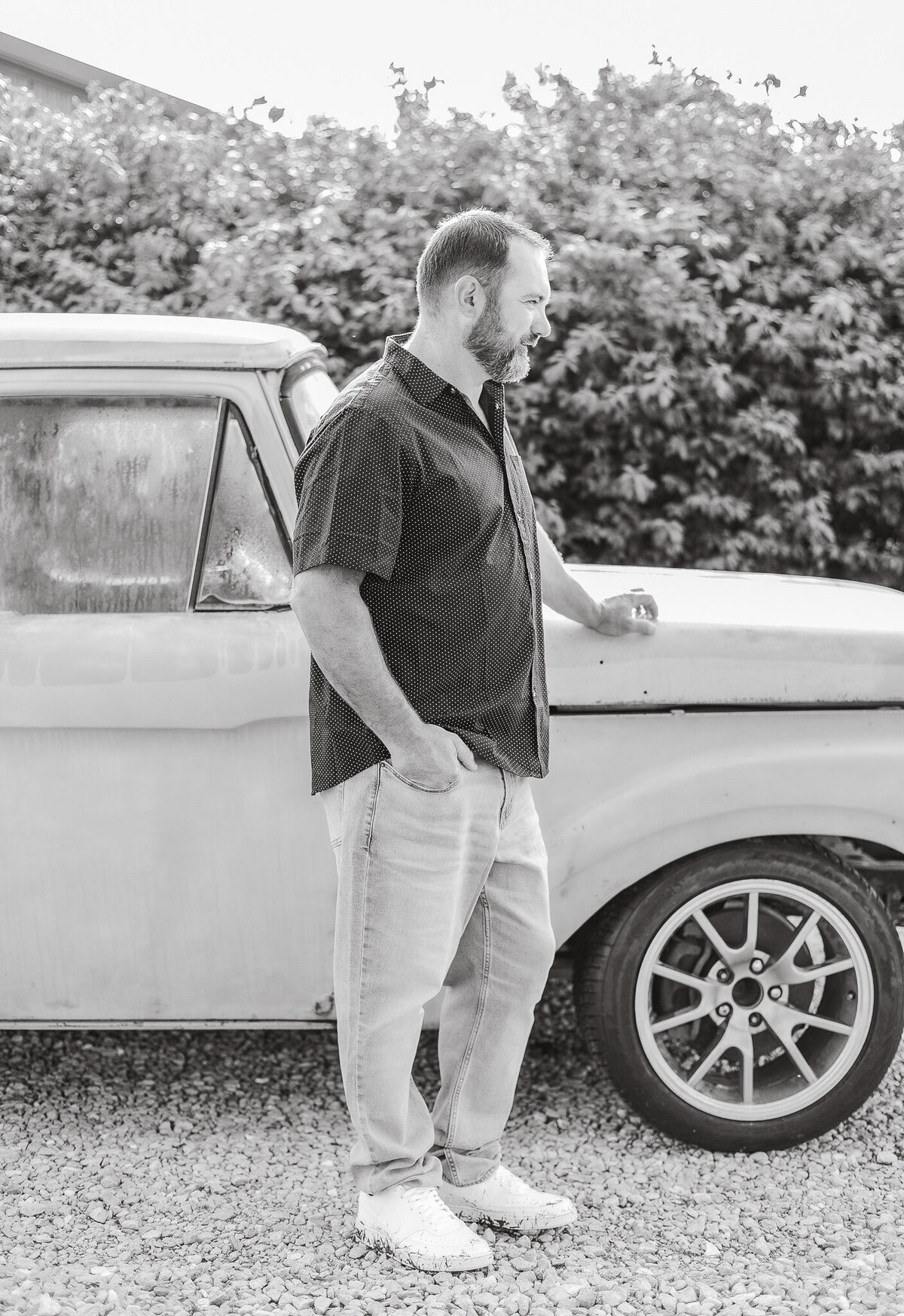 Black and white photo of Dad standing next to the classic truck, looking away from the camera, with a hand in his pocket.  Wearing blue jeans and short sleeve button up shirt.