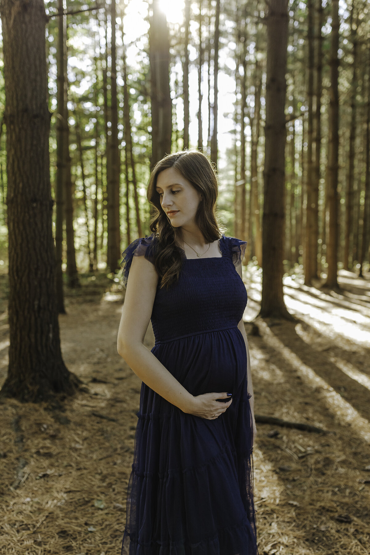Verona B Photography captures a gorgeous maternity picture of a mom holding her belly at Silver Creek metro Park in Norton, OH. The mom looks over her shoulder serenely with the sun peeking through the trees in this forest-vibe setting near Akron, OH.