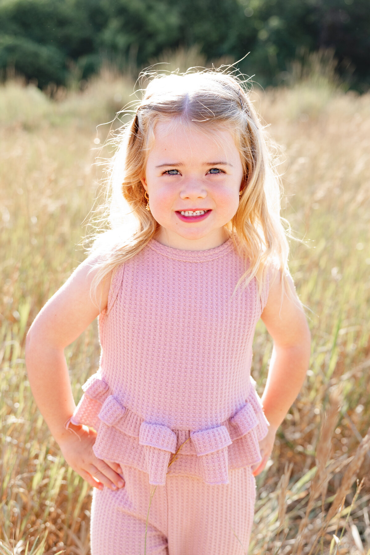 A young girl with blonde hair smiles brightly in a sunlit field, wearing a pink ruffled outfit. The golden hour light highlights her joyful expression.