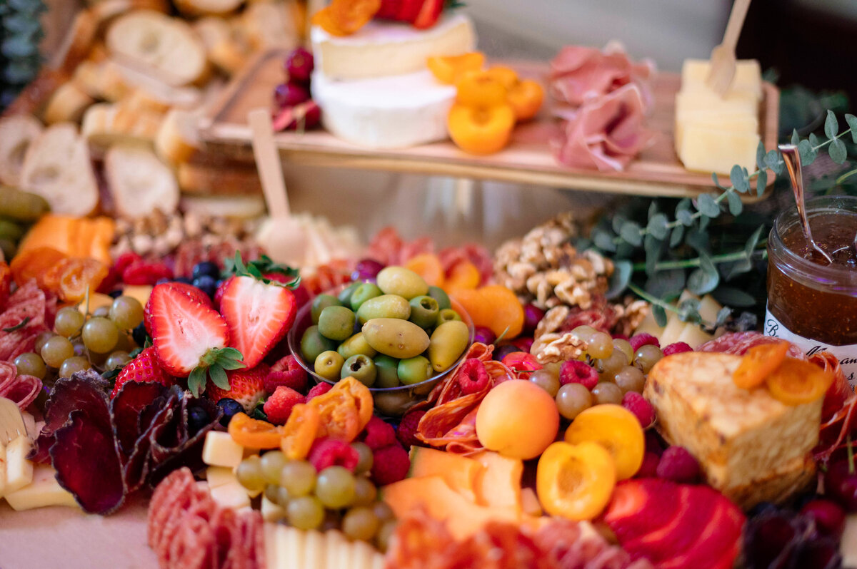 A vibrant charcuterie board filled with green olives, strawberries, grapes, cured meats, assorted cheeses, apricots, nuts, and bread, artistically arranged with greenery and a jar of jam.