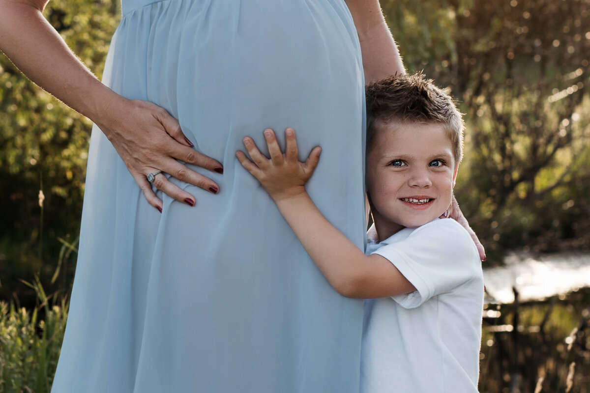6 year old son holding pregnant mom's belly who is wearing a blue maternity dress in a outdoor park
