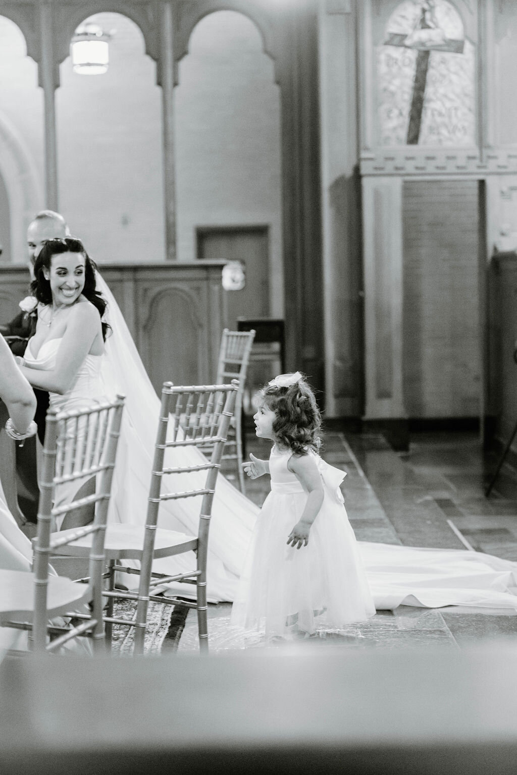 Rhode Island Wedding Photographers | A joyful bride in a gown and veil smiles at a young flower girl in a white dress walking down an aisle in a church, surrounded by chairs. Black and white photo.
