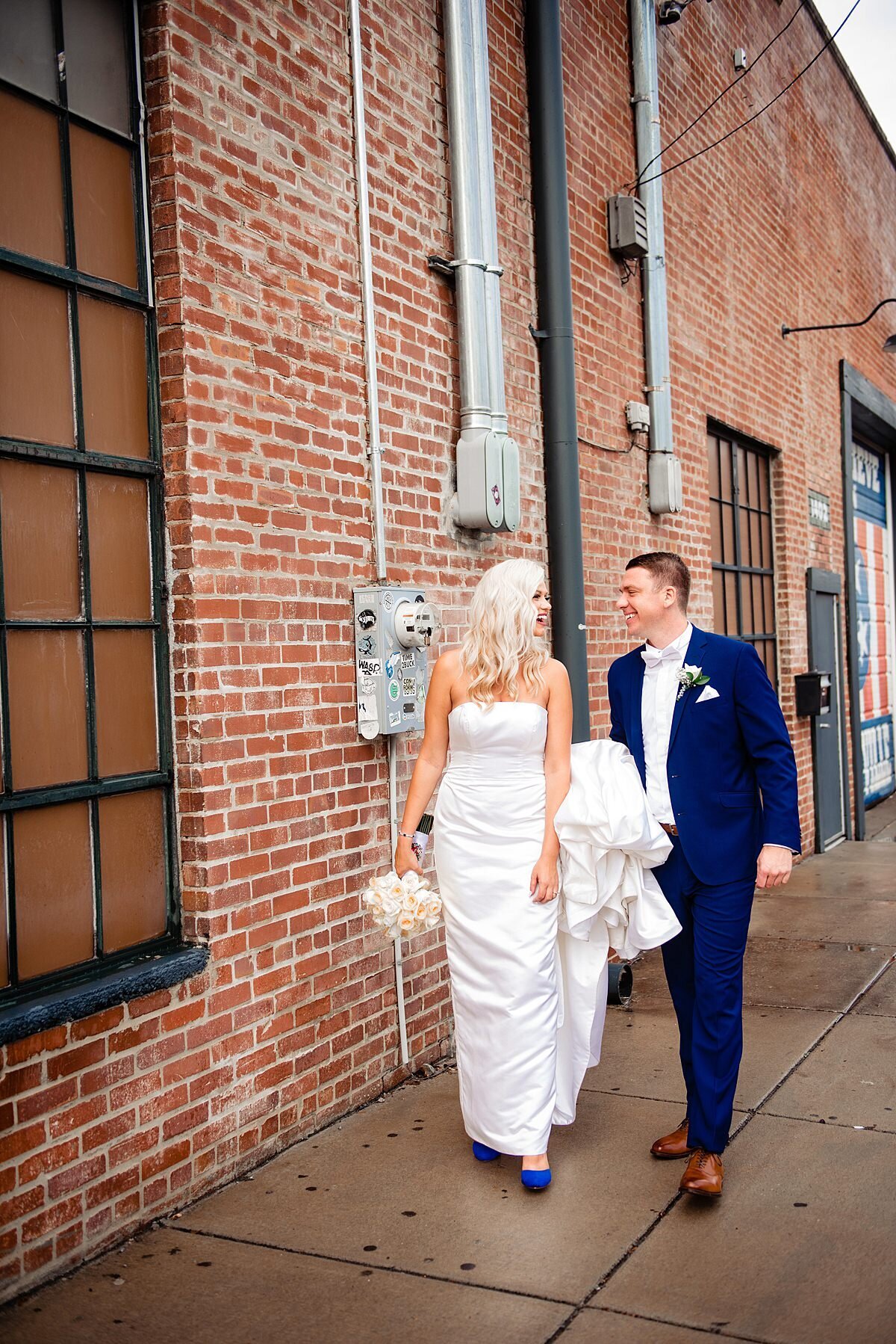 The groom carries the bride's train as they walk beside a brick building The bride and groom are looking at each other smiling. The bride is carrying a bouquet of white flowers.