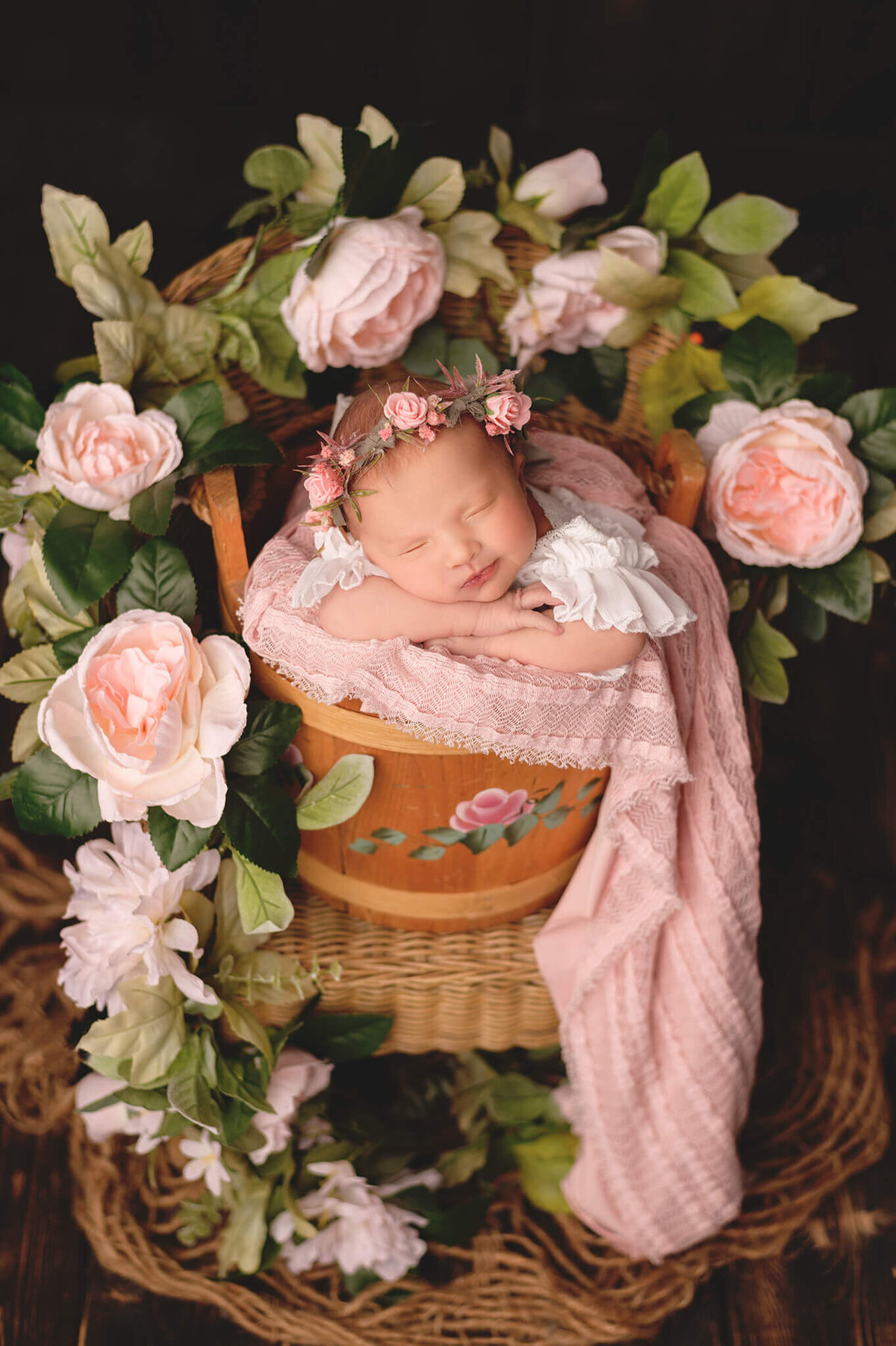 Newborn baby in a floral bucket surrounded by rose vines in a wicker chair at her studio session.