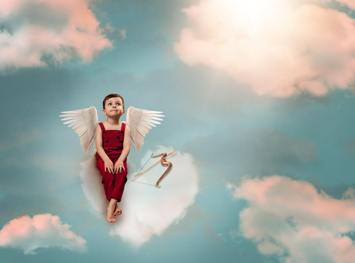 A little boy dressed as cupid sits on a cloud and rests