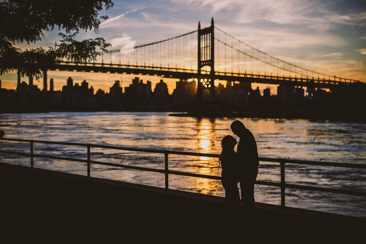 a silhouette of a couple having a moment at sunset at astoria park nyc with a bridge and river behind them